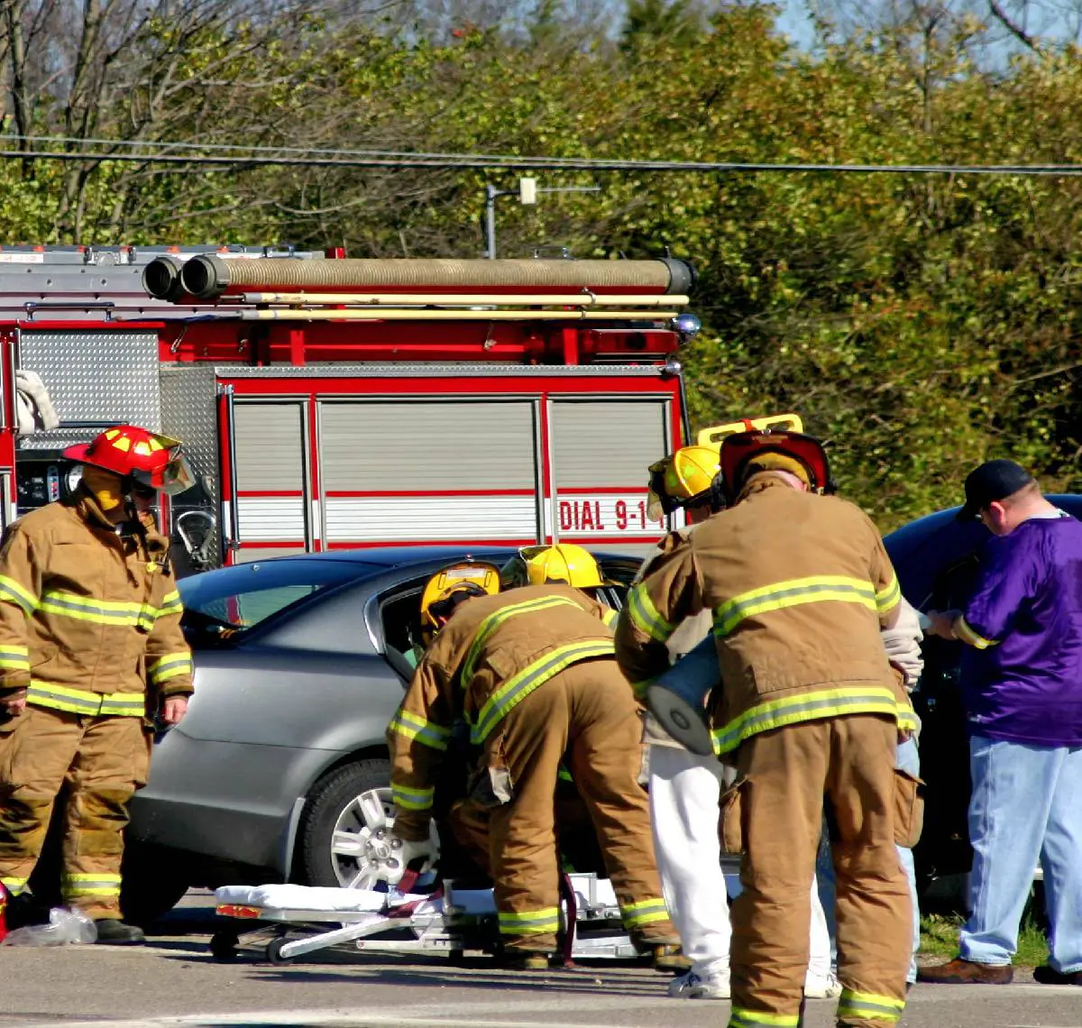 Firefighters attending to an emergency near a silver car with a fire truck in the background.