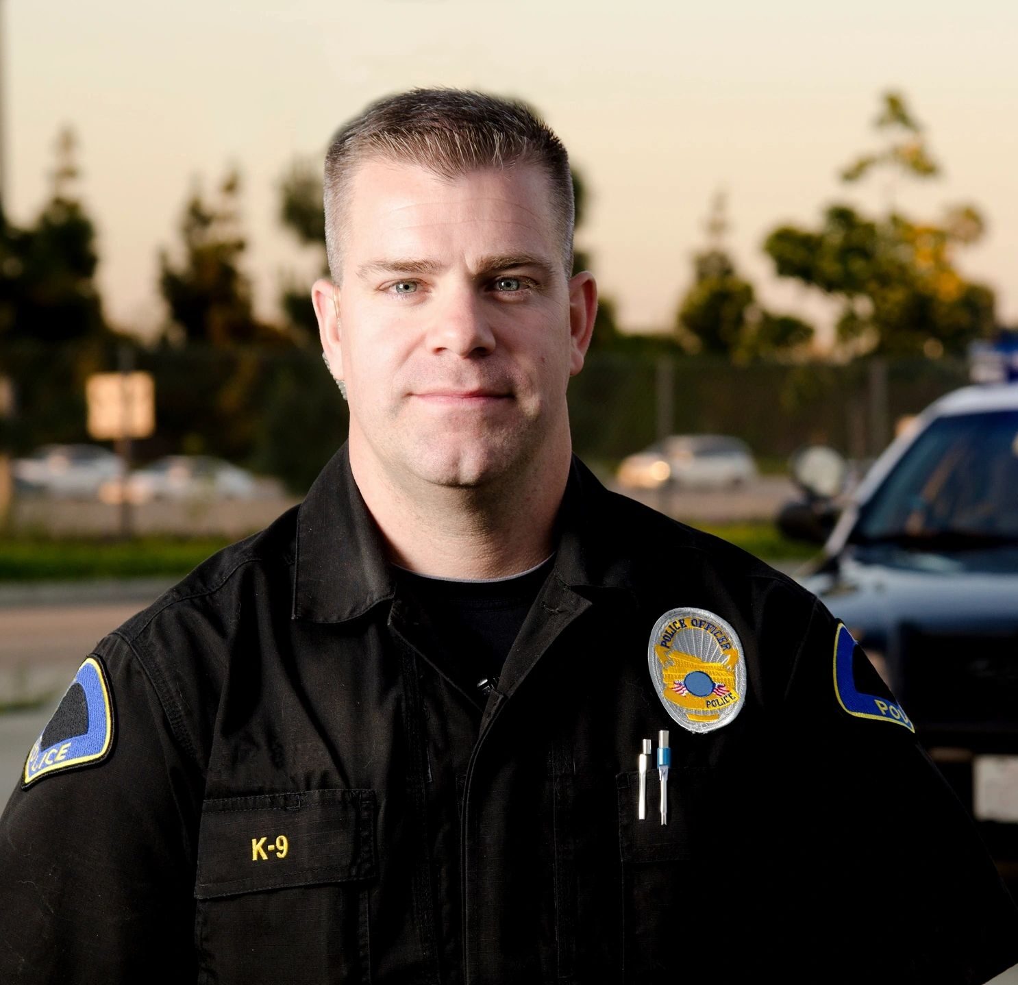Police officer standing outdoors with patrol cars and trees in the background.